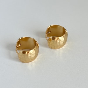 May include: A pair of gold-toned hoop earrings with a hammered texture. The earrings are thick and have a rounded shape.
