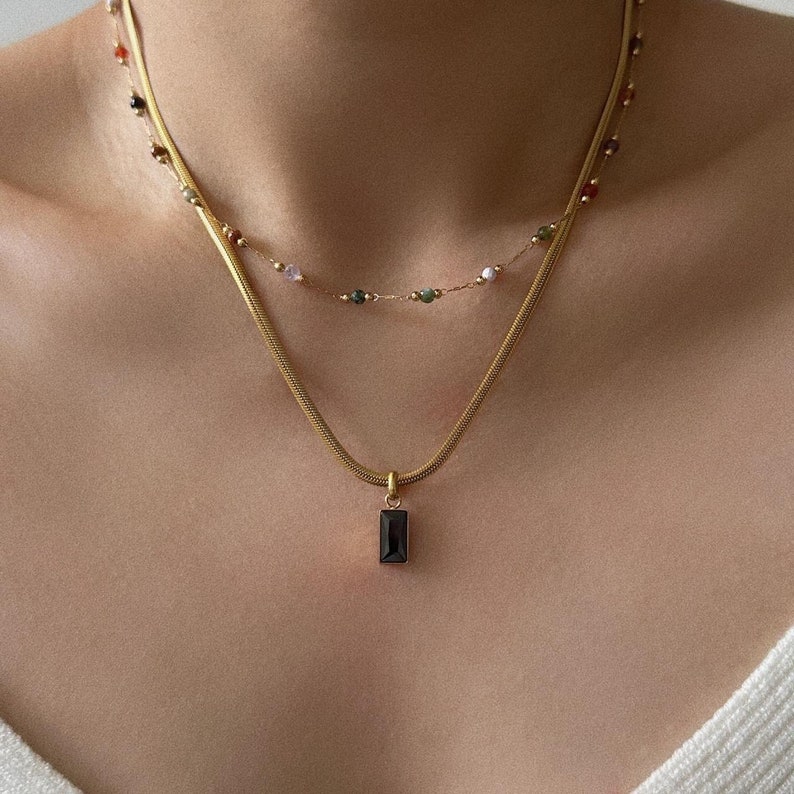 May include: A gold chain necklace with a black rectangular pendant. The chain is layered over a second chain with small, colourful beads.