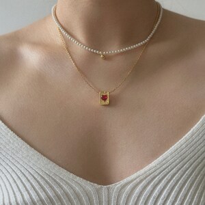 Peut inclure: Un collier en cha&icirc;ne dor&eacute;e avec un pendentif en forme de c&oelig;ur rouge. Le pendentif est serti dans un cadre carr&eacute; avec un motif textur&eacute;. Le collier est superpos&eacute; &agrave; un collier de perles avec une perle dor&eacute;e &agrave; l'extr&eacute;mit&eacute;.
