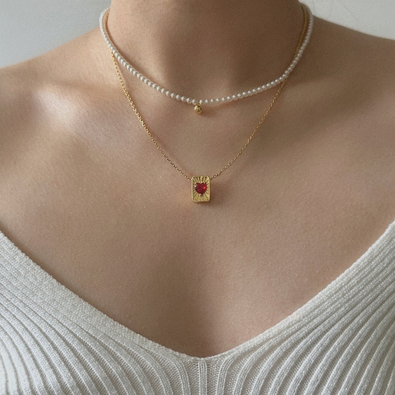 Peut inclure: Un collier en cha&icirc;ne dor&eacute;e avec un pendentif en forme de c&oelig;ur rouge. Le pendentif est serti dans un cadre carr&eacute; avec un motif textur&eacute;. Le collier est superpos&eacute; &agrave; un collier de perles avec une perle dor&eacute;e &agrave; l'extr&eacute;mit&eacute;.
