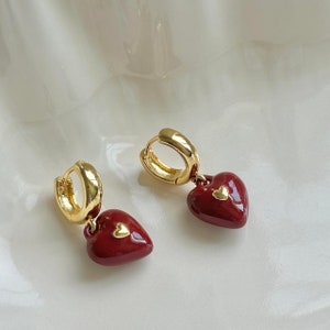 May include: A pair of gold hoop earrings with a dangling red heart charm. Each heart charm has a smaller gold heart in the center.
