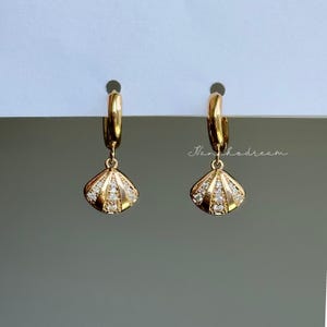 May include: A pair of gold-colored hoop earrings. Each earring features a small, fan-shaped charm adorned with clear, sparkling stones. The earrings are suspended from a gold hoop. The background is a light gray and white.