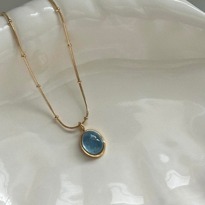 Peut inclure: Un collier dor&eacute; avec une cha&icirc;ne d&eacute;licate et un pendentif orn&eacute; d'une pierre pr&eacute;cieuse ovale bleu clair. Le pendentif est encadr&eacute; d'une lunette dor&eacute;e. Le collier est pr&eacute;sent&eacute; sur une surface blanche.