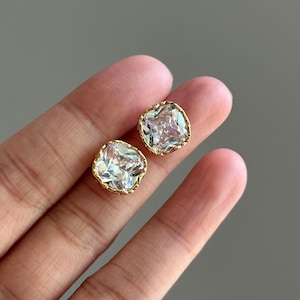 May include: A pair of square-shaped stud earrings. Each earring features a large, clear, faceted gemstone set in a gold-colored, textured bezel. The earrings are displayed against a neutral background.