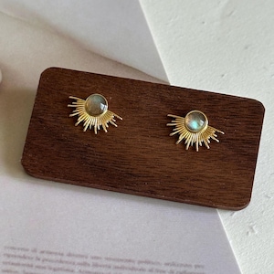 May include: A pair of gold-coloured stud earrings. Each earring has a round, iridescent stone at the centre, surrounded by a sunburst design. The earrings are displayed on a dark brown wooden surface.