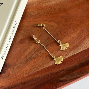 May include: A pair of gold earrings with a ginkgo leaf design. The earrings are dangling and have a delicate chain.
