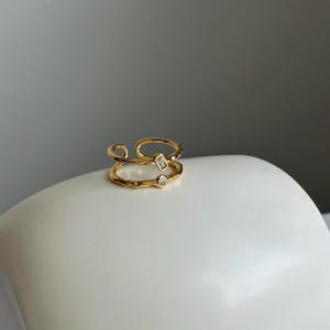 May include: A gold ring with two clear stones. The ring is made of a textured metal and has a unique design.