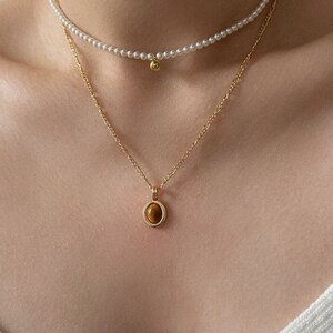 Peut inclure: Un collier en cha&icirc;ne dor&eacute;e avec un pendentif en &oelig;il de tigre. Le pendentif est serti dans un cercle d'or avec un cercle d'or plus petit autour. Le collier est superpos&eacute; &agrave; un collier de perles avec un petit charme en or.