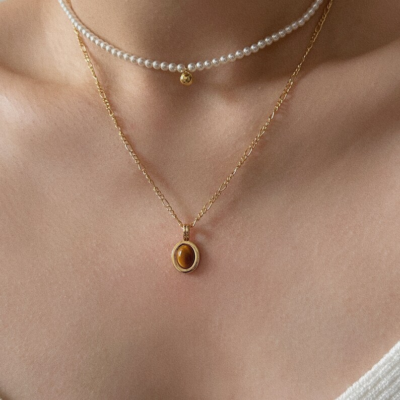 Peut inclure: Un collier en cha&icirc;ne dor&eacute;e avec un pendentif en &oelig;il de tigre. Le pendentif est serti dans un cercle d'or avec un cercle d'or plus petit autour. Le collier est superpos&eacute; &agrave; un collier de perles avec un petit charme en or.