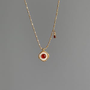 May include: A gold-toned necklace with a pendant featuring a round, red gemstone set in a white and gold frame. A small, teardrop-shaped red gemstone hangs from the chain. The necklace is displayed against a gray background.