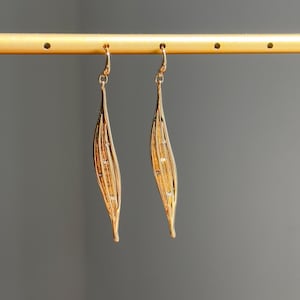 May include: A pair of gold-colored dangle earrings. Each earring has a long, leaf-like shape with delicate lines and small, sparkling accents. The earrings are suspended from a gold-colored hook.