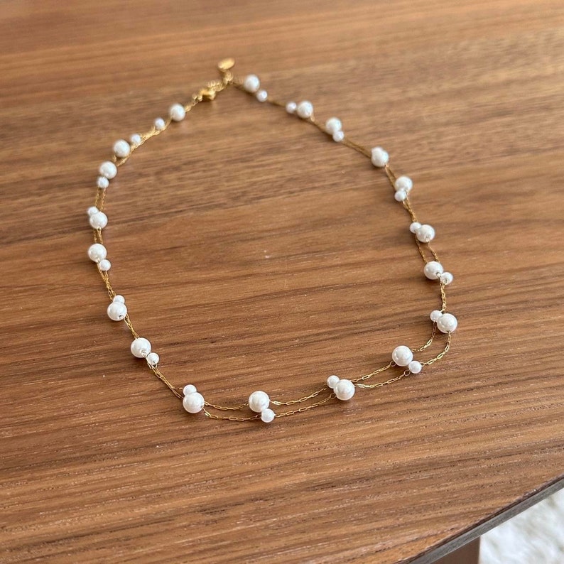 K&ouml;nnte beinhalten: Eine zarte goldene Kettenkette mit kleinen wei&szlig;en Perlen. Die Kette ist doppelf&auml;dig und hat ein schlichtes Design.