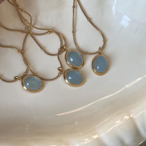 Peut inclure: Plusieurs colliers dor&eacute;s orn&eacute;s de pendentifs ovales bleu clair. Chaque pendentif, encadr&eacute; d'or, est suspendu &agrave; une cha&icirc;ne d&eacute;licate. Les colliers sont pr&eacute;sent&eacute;s sur une surface blanche textur&eacute;e.