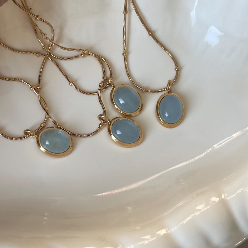 Peut inclure: Plusieurs colliers dor&eacute;s orn&eacute;s de pendentifs ovales bleu clair. Chaque pendentif, encadr&eacute; d'or, est suspendu &agrave; une cha&icirc;ne d&eacute;licate. Les colliers sont pr&eacute;sent&eacute;s sur une surface blanche textur&eacute;e.
