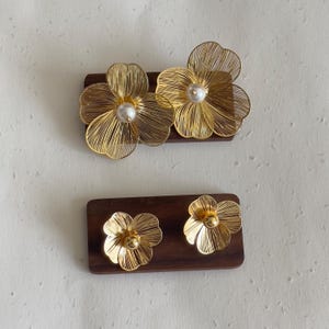 May include: Gold-colored floral earrings and a matching brooch set on a dark wooden base. The earrings and brooch feature delicate wire-like petals and a pearl-like center. The set is displayed against a neutral background.