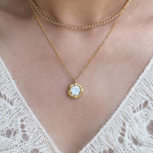 May include: A gold necklace with a pendant holding a white, irregularly shaped stone. The pendant has a textured gold frame. A second, delicate gold chain with small, clear stones is also visible.