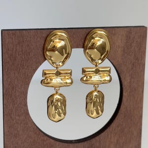 May include: A pair of gold-tone dangle earrings with a textured, abstract design. The earrings feature three tiers of geometric shapes.