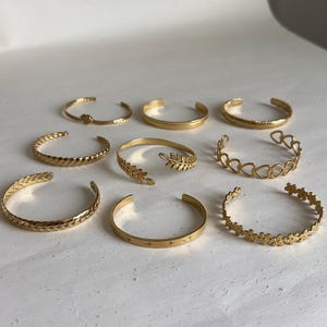 May include: Nine gold-coloured open cuff bracelets. The bracelets display diverse designs, including braided, leaf, heart, and floral patterns. Some have simple, smooth bands, while others have textured surfaces.