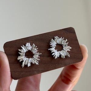 May include: Two silver-colored stud earrings with a radial, spiky design, resembling a burst of light. The earrings are displayed on a small, rectangular, dark wooden surface. The background is a neutral, light color.