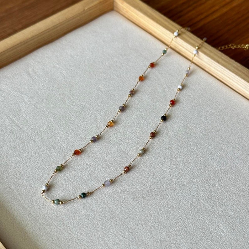 May include: A gold chain necklace with small, colourful gemstone beads. The beads are various shades of red, orange, green, purple, and black.