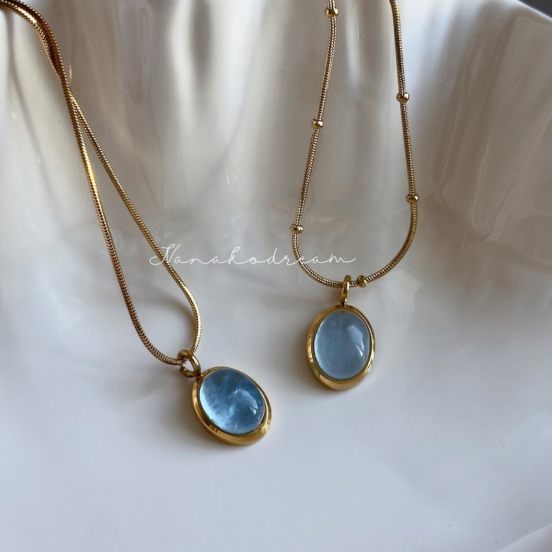 Peut inclure: Deux colliers dor&eacute;s avec des pendentifs ovales bleu clair. Les pendentifs sont sertis dans des montures dor&eacute;es. Les colliers sont pr&eacute;sent&eacute;s sur une surface blanche textur&eacute;e. Le texte "Nanahodream" est &eacute;crit en cursive.