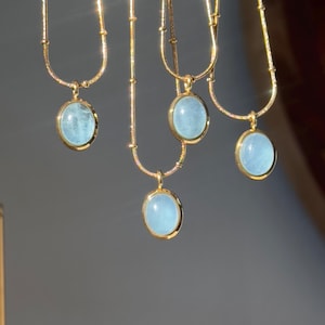 Peut inclure: Plusieurs colliers dor&eacute;s orn&eacute;s de pendentifs ovales bleu clair. Les pendentifs sont sertis dans des cadres dor&eacute;s. Les colliers sont suspendus sur un fond gris neutre, la lumi&egrave;re se refl&eacute;tant sur les cha&icirc;nes et les pendentifs dor&eacute;s.