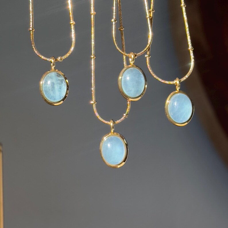 Peut inclure: Plusieurs colliers dor&eacute;s orn&eacute;s de pendentifs ovales bleu clair. Les pendentifs sont sertis dans des cadres dor&eacute;s. Les colliers sont suspendus sur un fond gris neutre, la lumi&egrave;re se refl&eacute;tant sur les cha&icirc;nes et les pendentifs dor&eacute;s.