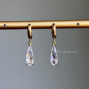 May include: A pair of gold-colored hoop earrings with teardrop-shaped, clear crystal pendants. The earrings are hanging from a gold bar against a gray background. The crystals reflect light, creating a sparkling effect.