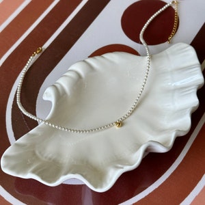 Peut inclure: Un collier d&eacute;licat de perles avec une seule perle dor&eacute;e &agrave; l'extr&eacute;mit&eacute;. Le collier est pr&eacute;sent&eacute; sur un plat blanc en forme de coquille.