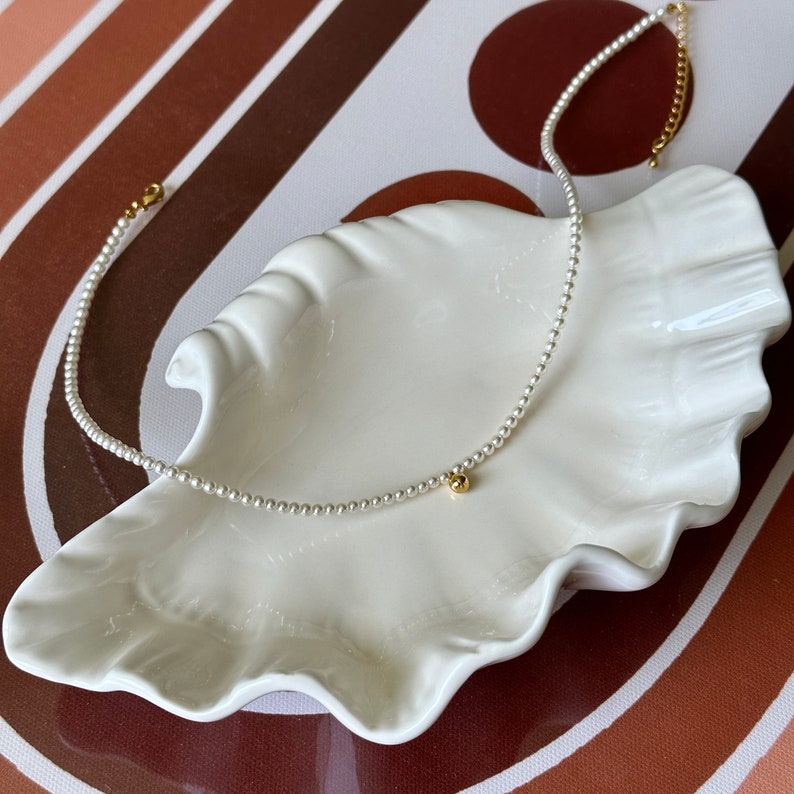 Peut inclure: Un collier d&eacute;licat de perles avec une seule perle dor&eacute;e &agrave; l'extr&eacute;mit&eacute;. Le collier est pr&eacute;sent&eacute; sur un plat blanc en forme de coquille.