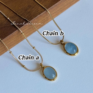 Peut inclure: Deux colliers dor&eacute;s, &eacute;tiquet&eacute;s "Cha&icirc;ne A" et "Cha&icirc;ne B". Chaque collier pr&eacute;sente une cha&icirc;ne fine et un pendentif avec une pierre ovale bleu clair sertie dans un cadre dor&eacute;. L'arri&egrave;re-plan est de couleur claire.
