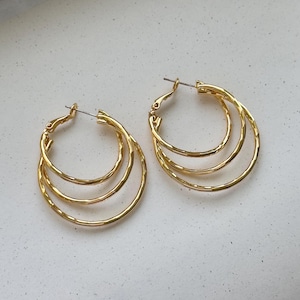 May include: A pair of gold-colored hoop earrings. Each earring features three nested hoops with a textured surface. The earrings have a classic, elegant design and are suitable for everyday wear or special occasions.