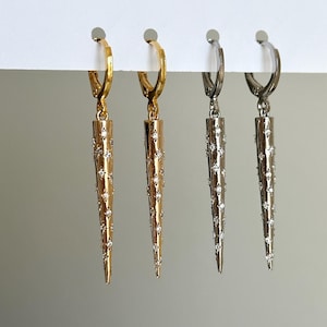 May include: Three pairs of earrings with a spike design. The earrings are made of gold, silver, and black metal. Each spike is covered in small, clear rhinestones.
