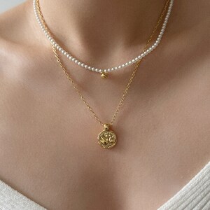 Peut inclure: Un collier en cha&icirc;ne dor&eacute;e avec un pendentif rond orn&eacute; d'un motif floral. Le collier est superpos&eacute; &agrave; un collier de perles avec une petite boule dor&eacute;e &agrave; l'extr&eacute;mit&eacute;.