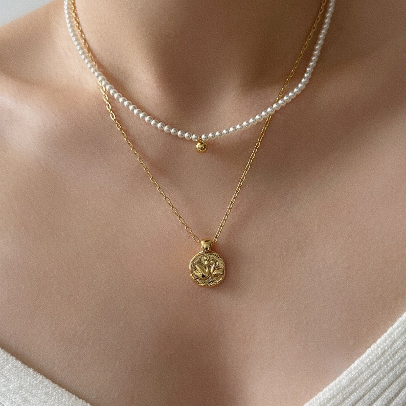 Peut inclure: Un collier en cha&icirc;ne dor&eacute;e avec un pendentif rond orn&eacute; d'un motif floral. Le collier est superpos&eacute; &agrave; un collier de perles avec une petite boule dor&eacute;e &agrave; l'extr&eacute;mit&eacute;.