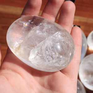 May include: A clear quartz crystal palm stone with a smooth, polished surface. The stone is held in a hand, and the light reflects off the crystal, creating a rainbow effect.