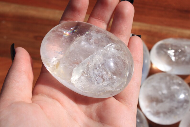May include: A clear quartz crystal palm stone with a smooth, polished surface. The stone is held in a hand, and the light reflects off the crystal, creating a rainbow effect.