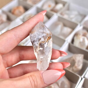 May include: A clear quartz crystal with a pointed tip, held in a hand. The crystal has a rough, textured surface with areas of white and brown. Other crystals are visible in the background, stored in a divided box.