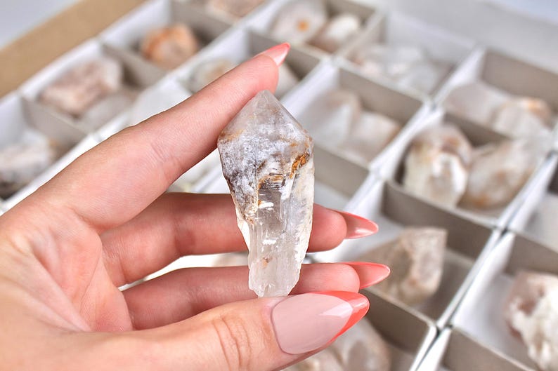 May include: A clear quartz crystal with a pointed tip, held in a hand. The crystal has a rough, textured surface with areas of white and brown. Other crystals are visible in the background, stored in a divided box.