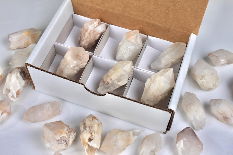 May include: A white cardboard box with a brown interior holds several raw quartz crystals. The crystals are various sizes and shapes, with a mix of clear and beige tones. Some crystals are scattered around the box.