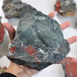 May include: A large, rough-hewn stone with a mix of dark green, gray, and red hues. The stone is held in a hand, with several other similar stones scattered around. The overall appearance suggests a natural, raw mineral.