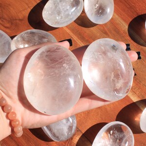 May include: A collection of clear quartz crystal stones, some of which are smooth and polished, while others are more rough and natural. The stones are arranged on a wooden surface, and one is being held in a hand wearing a bracelet.