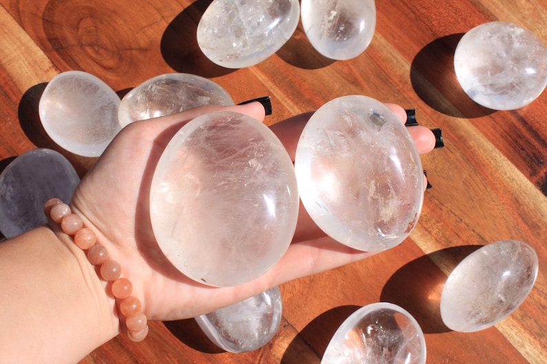 May include: A collection of clear quartz crystal stones, some of which are smooth and polished, while others are more rough and natural. The stones are arranged on a wooden surface, and one is being held in a hand wearing a bracelet.