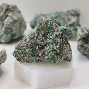 May include: A cluster of green emerald crystals embedded in a gray rock. The rock is sitting on a white pedestal.