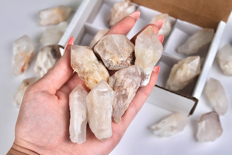 May include: A collection of raw quartz crystals in varying shapes and sizes. Some crystals are held in a hand, while others are arranged in a white box and scattered on a white surface. The crystals display a range of colors, from clear to milky white and light brown.