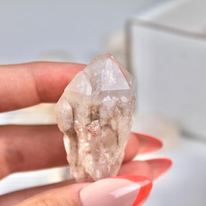 May include: A clear quartz crystal with a pointed tip and a rough, textured base. The crystal has a light, translucent appearance with subtle variations in color, ranging from clear to a pale beige. The crystal is held in a hand.