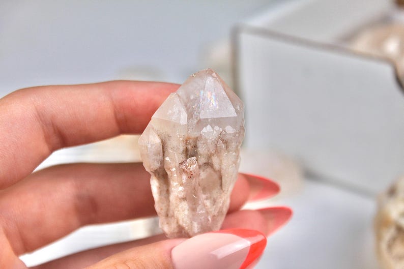 May include: A clear quartz crystal with a pointed tip and a rough, textured base. The crystal has a light, translucent appearance with subtle variations in color, ranging from clear to a pale beige. The crystal is held in a hand.