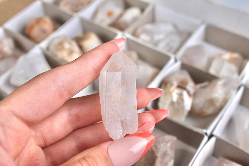 May include: A clear quartz crystal point is held in a hand, showcasing its natural, translucent appearance. The crystal is a light, neutral color, with a pointed tip and a rough base. Other crystals are visible in the background, stored in a divided box.
