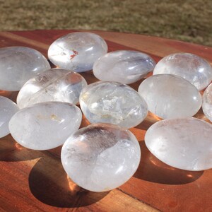 May include: A collection of 10 smooth, clear quartz crystal stones, each shaped like a rounded oval, arranged in a starburst pattern on a wooden surface.