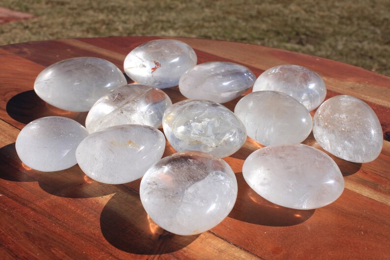 May include: A collection of 10 smooth, clear quartz crystal stones, each shaped like a rounded oval, arranged in a starburst pattern on a wooden surface.